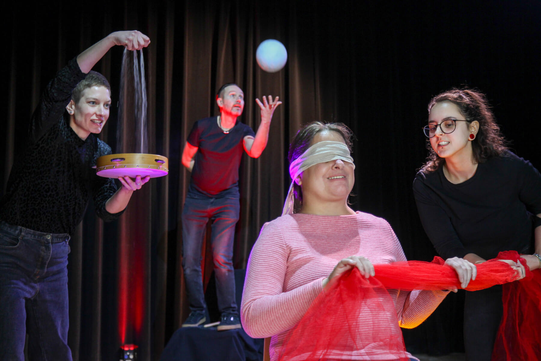 Theaterszene: Person mit verbundenen Augen wird von drei Performerinnen mit Geräuschen und Bewegungen umgeben.